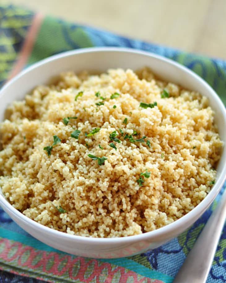 Bowl of cooked couscous garnished with herbs on a colorful patterned cloth, next to a spoon.