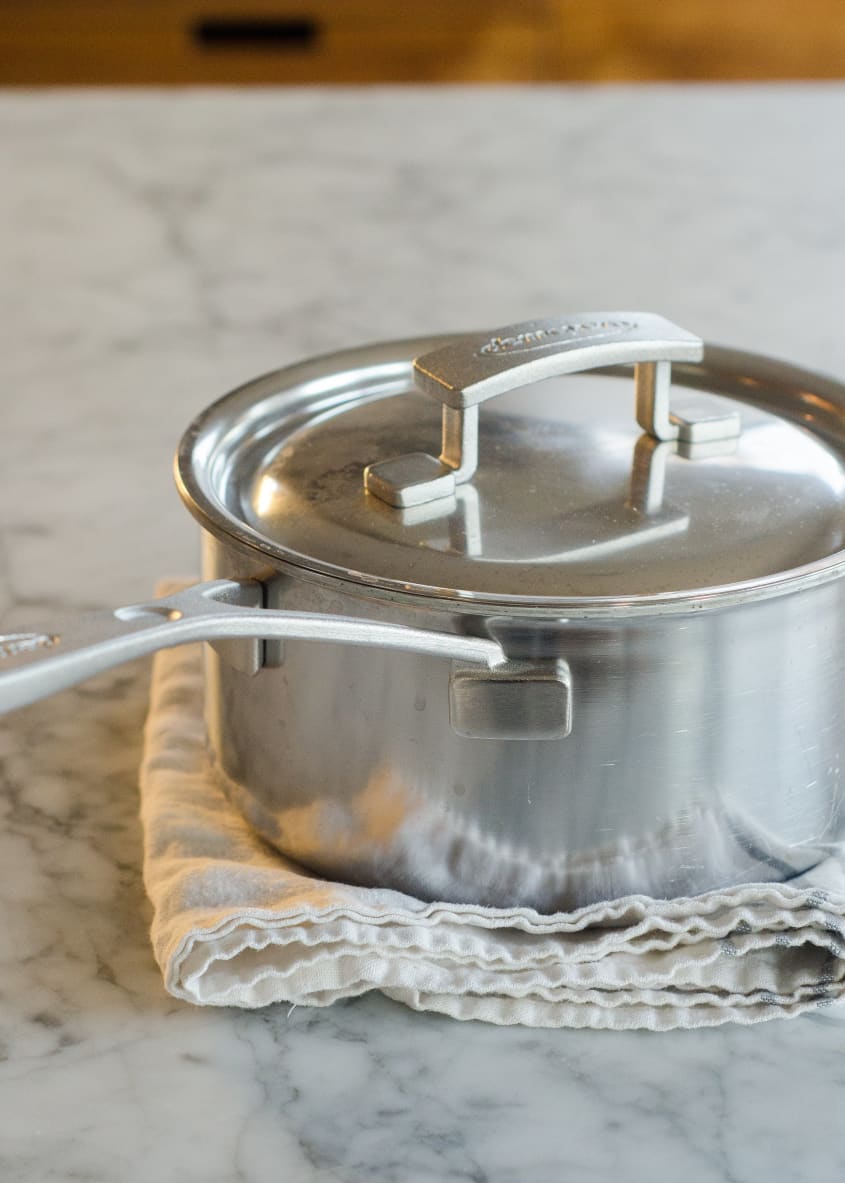 A stainless steel pot with a lid on a folded cloth, set on a marble countertop.