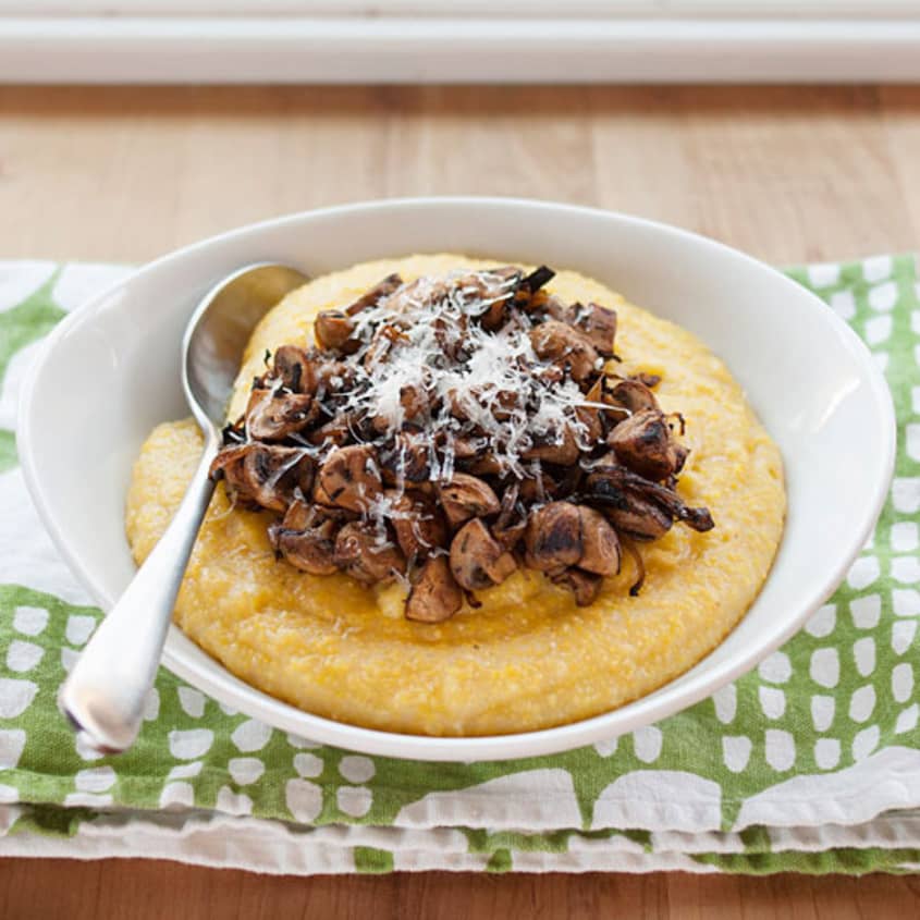 Creamy polenta topped with sautéed mushrooms and grated cheese in a white bowl with a spoon.