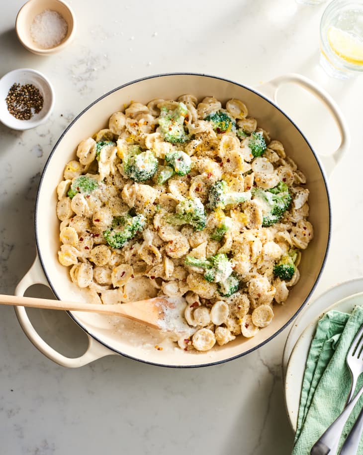 Orecchiette pasta with broccoli and creamy sauce in a large pot, garnished with pepper and cheese, on a marble surface.