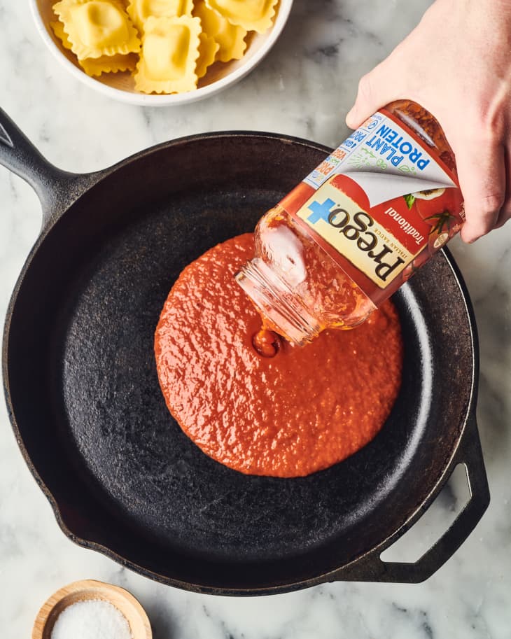 Pouring tomato sauce into a skillet with ravioli in a bowl nearby.