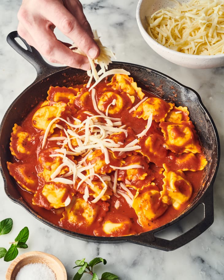 Ravioli in tomato sauce topped with shredded cheese in a skillet, garnished with fresh herbs.