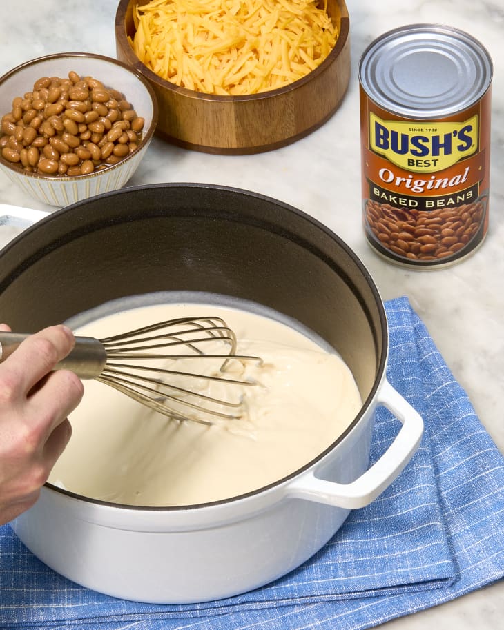 Whisking cheese sauce in a pot with Bush's baked beans and shredded cheese nearby.