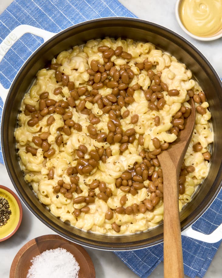 Mac and cheese topped with baked beans in a pot, wooden spoon, and side condiments.
