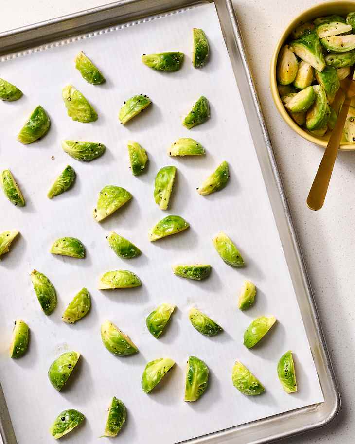 Brussels sprouts cut into wedges, seasoned, and arranged on a baking sheet with a bowl of mixed sprouts nearby.