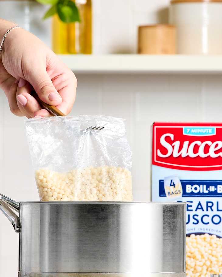 A hand holding a fork above a pot with a bag of pearl couscous ready to be cooked.