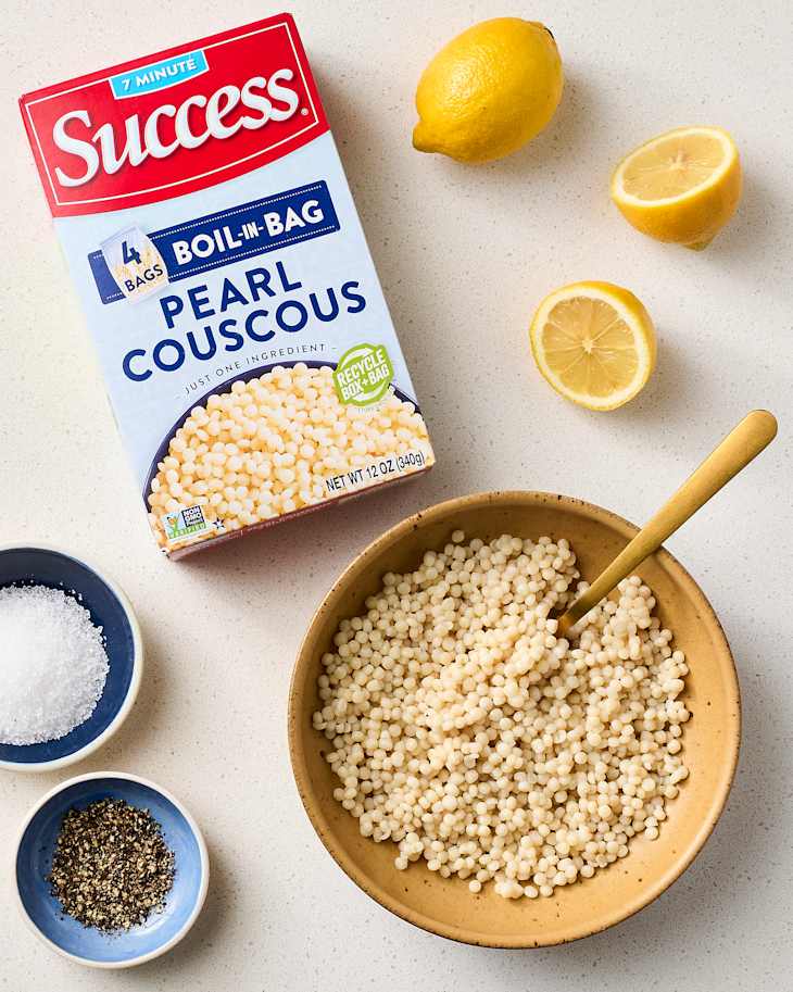A bowl of cooked pearl couscous with a wooden spoon, surrounded by lemon halves and bowls of salt and pepper.
