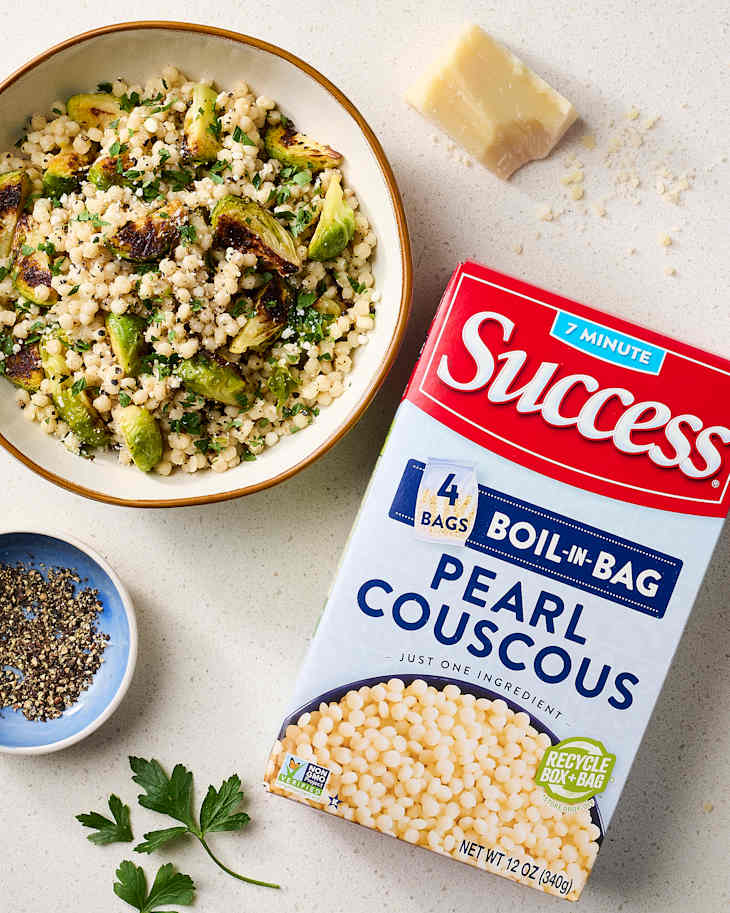 Bowl of pearl couscous mixed with Brussels sprouts and parsley, with a box of couscous and Parmesan nearby.
