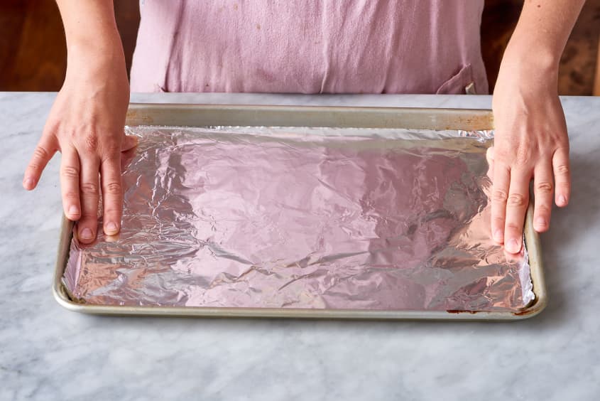 Hands lining a baking sheet with aluminum foil on a marble countertop.