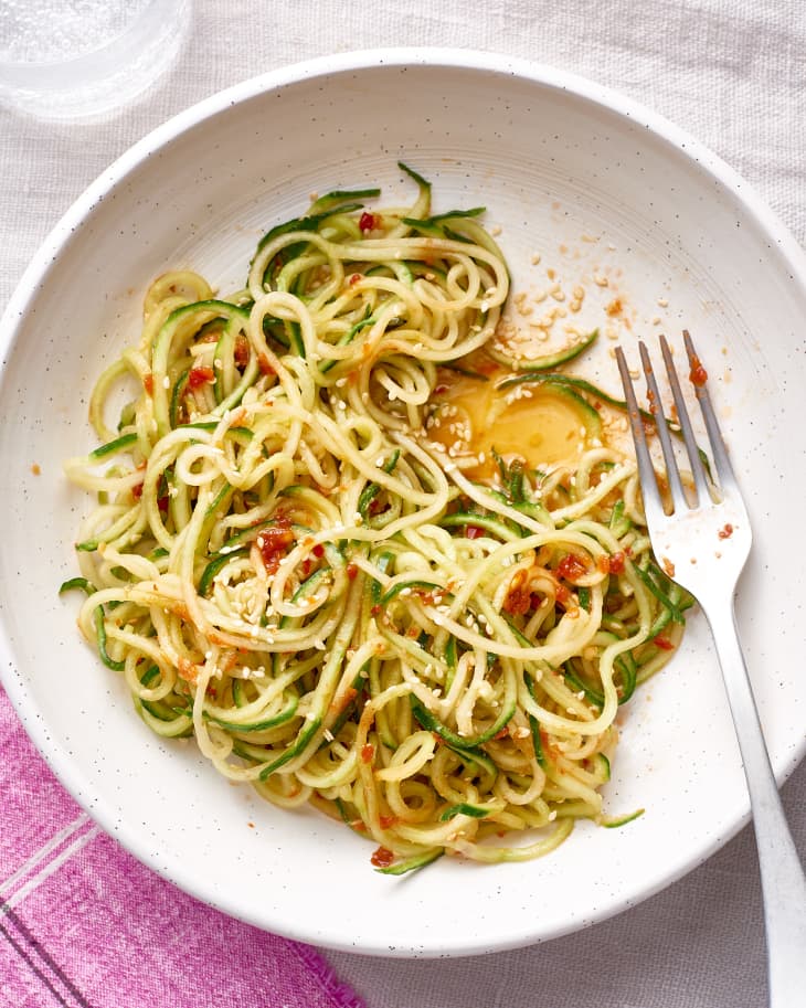 Cucumber noodles with chili-garlic sauce and sesame seeds in a white bowl, fork on the side.