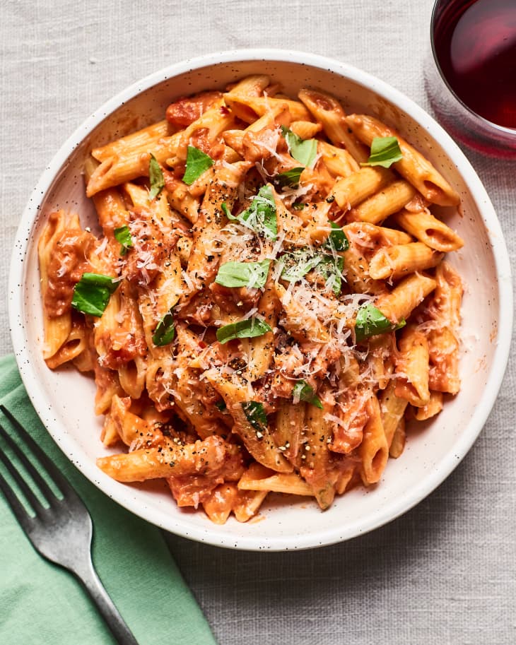 Creamy tomato pasta with penne, fresh basil, and grated cheese in a bowl, alongside a glass of red wine.