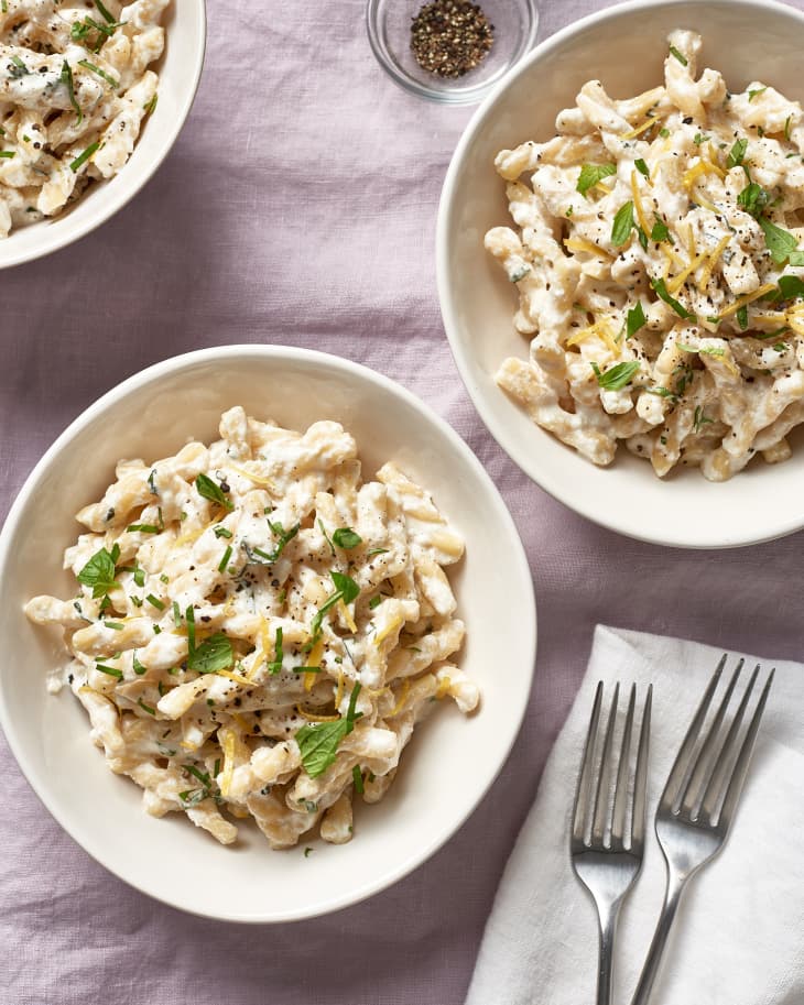Creamy pasta with herbs and lemon zest in white bowls, garnished with black pepper, next to two forks on a napkin.