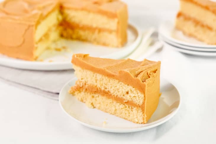 Slice of caramel cake with creamy frosting on a white plate, whole cake in background.