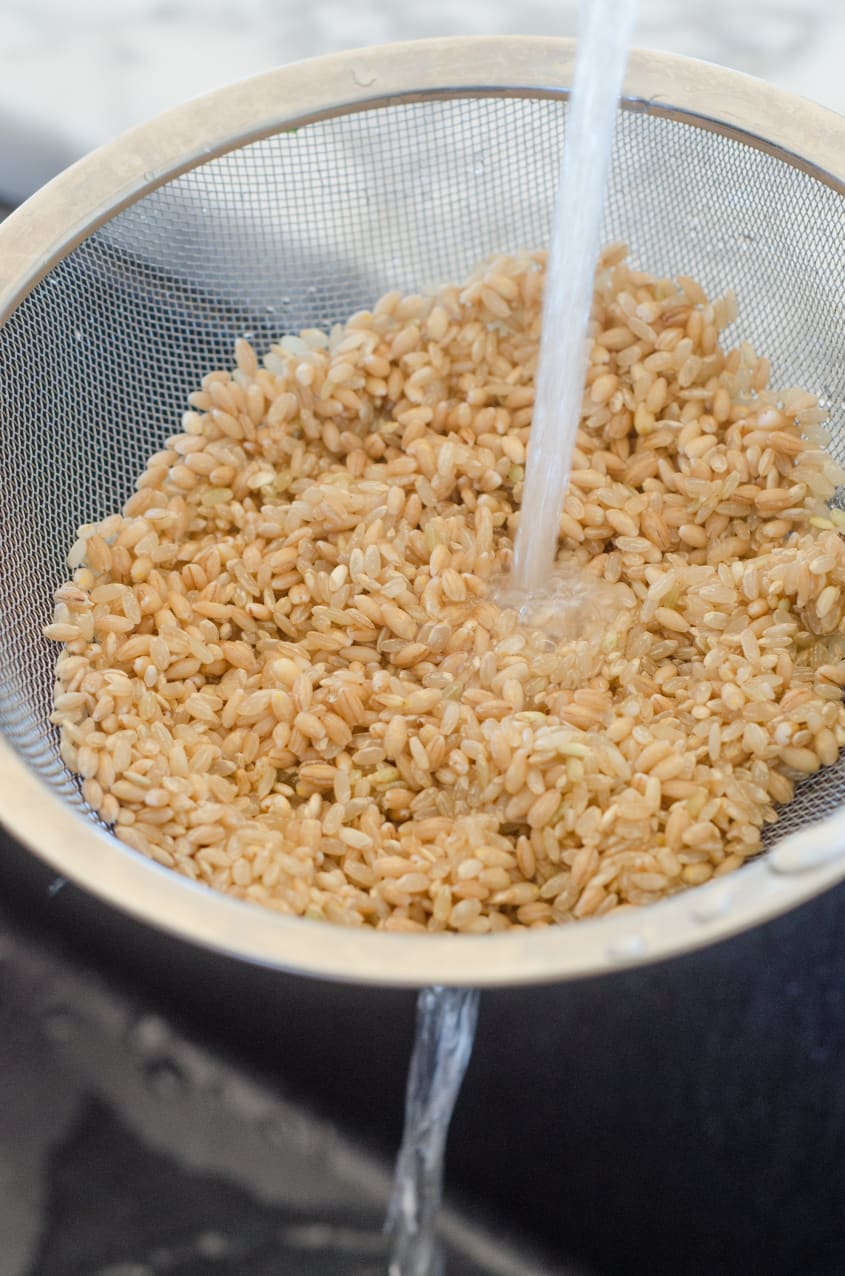 Brown rice being rinsed in a metal strainer under running water.