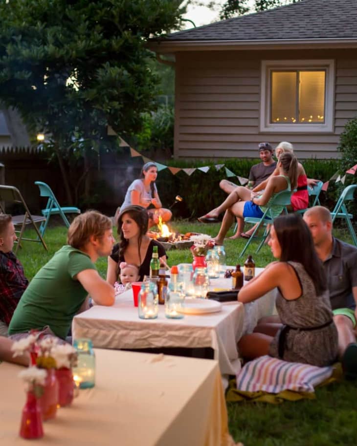 Backyard gathering with people sitting around tables and a fire pit, surrounded by string lights and colorful chairs.