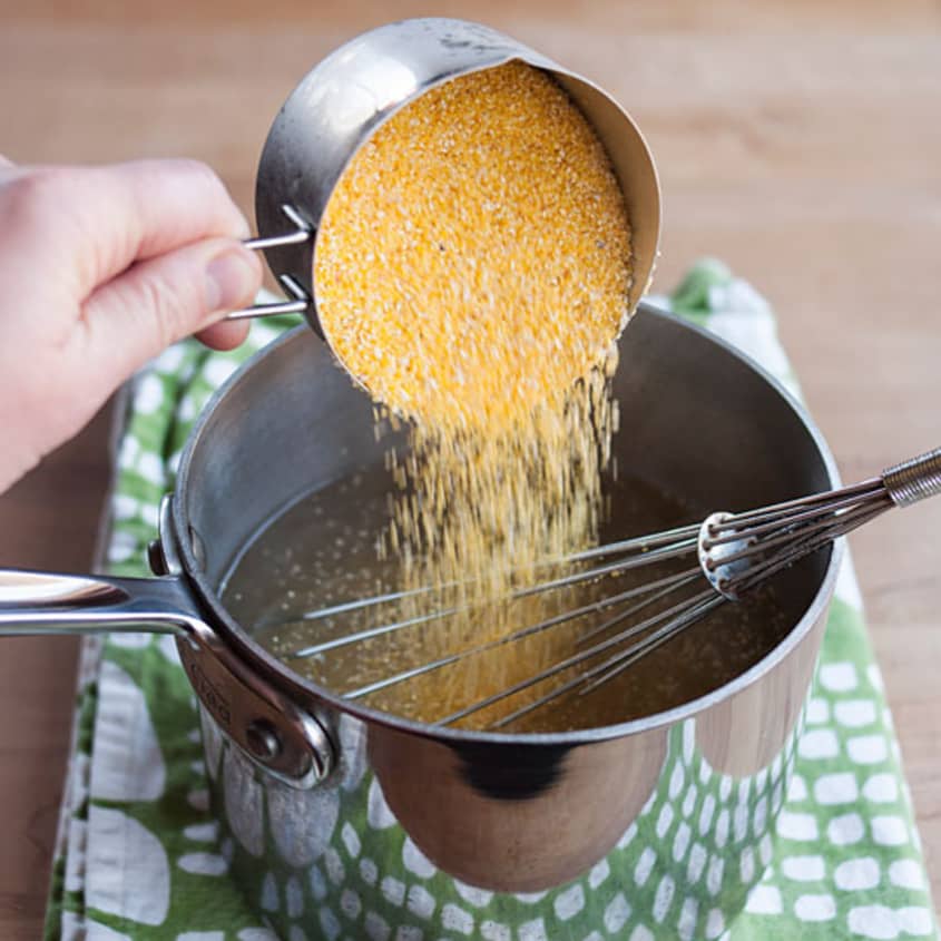 Pouring cornmeal into a saucepan with a whisk on a green towel.