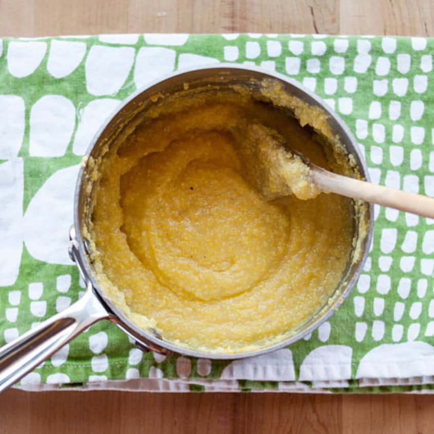 Pot of creamy polenta with a wooden spoon on a green patterned cloth.