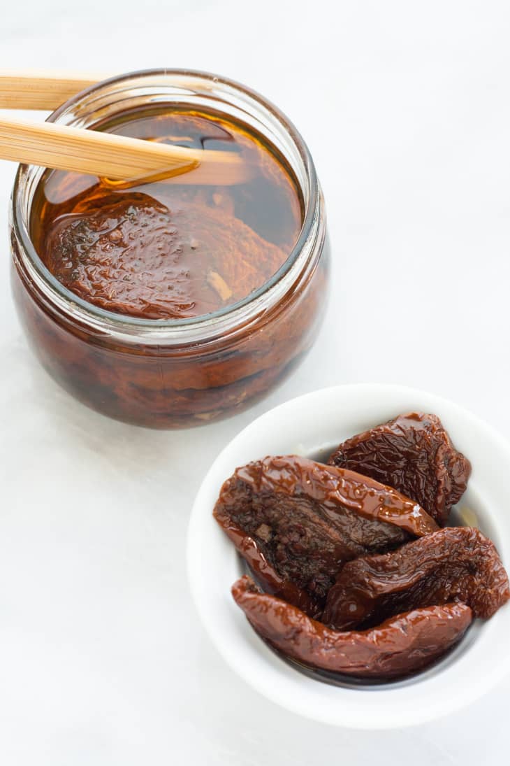 Sun-dried tomatoes in oil in a glass jar with wooden tongs, next to a small white bowl of sun-dried tomatoes.