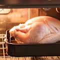 Raw turkey in a black roasting pan inside an oven, with visible oven racks and warm lighting.