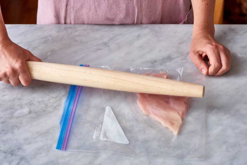 Person flattening chicken breast with a rolling pin inside a plastic bag on a marble countertop.