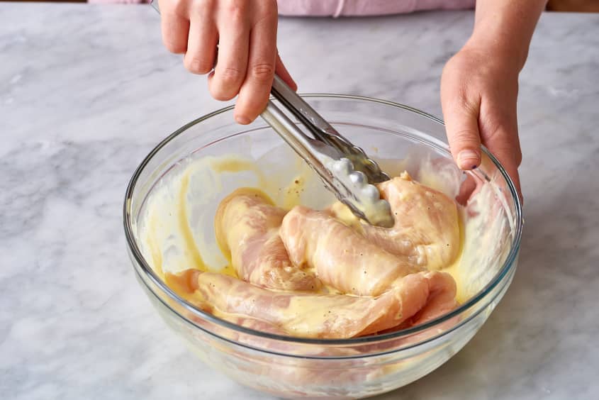 Raw chicken breasts being marinated in a glass bowl with tongs.