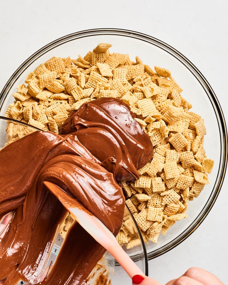 Mixing melted chocolate with cereal squares in a glass bowl using a spatula.