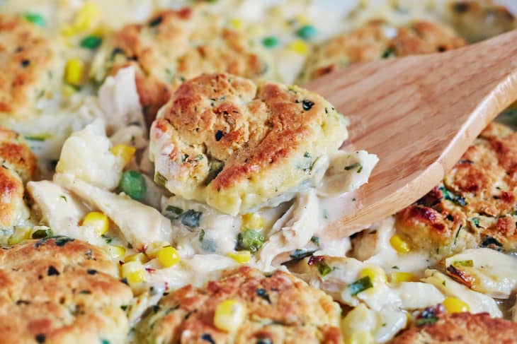 Chicken pot pie with golden biscuits, corn, peas, and creamy sauce being served with a wooden spoon.