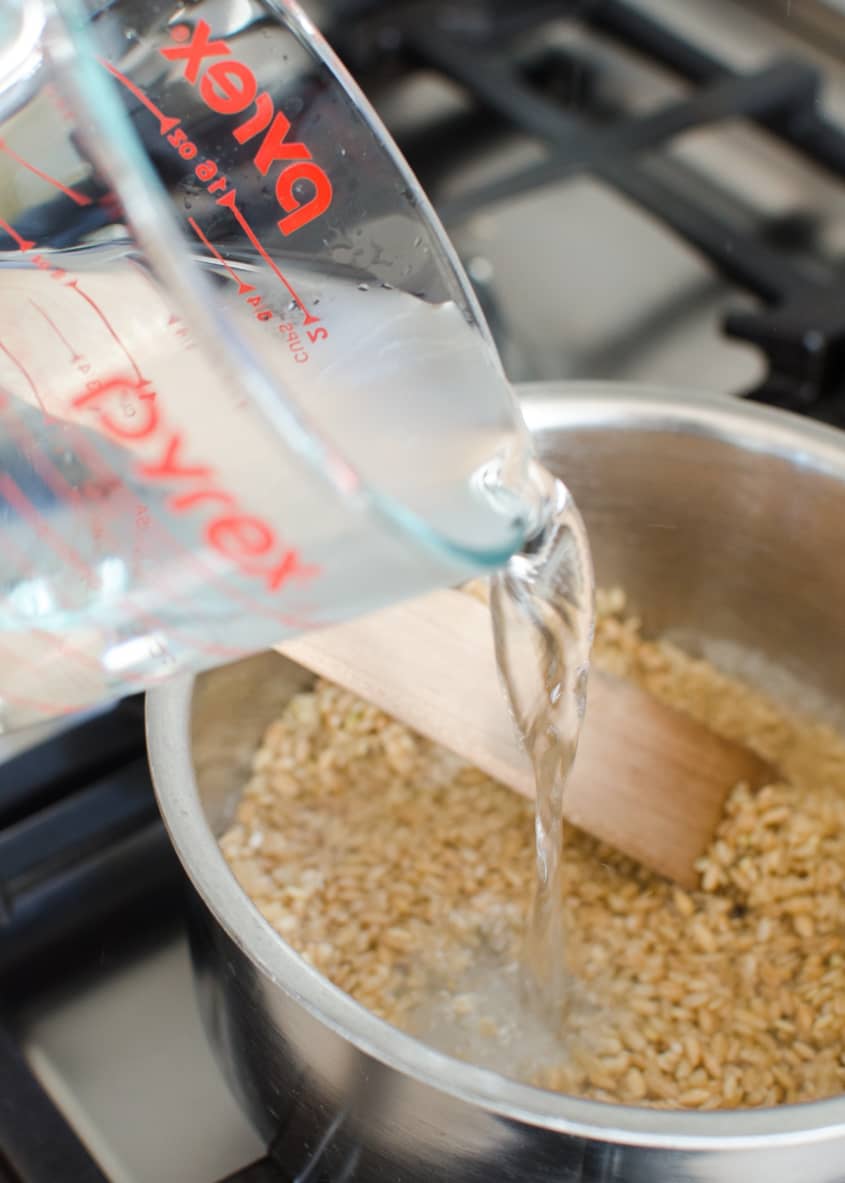 Pouring water from a measuring cup into a pot of brown rice on a stove.