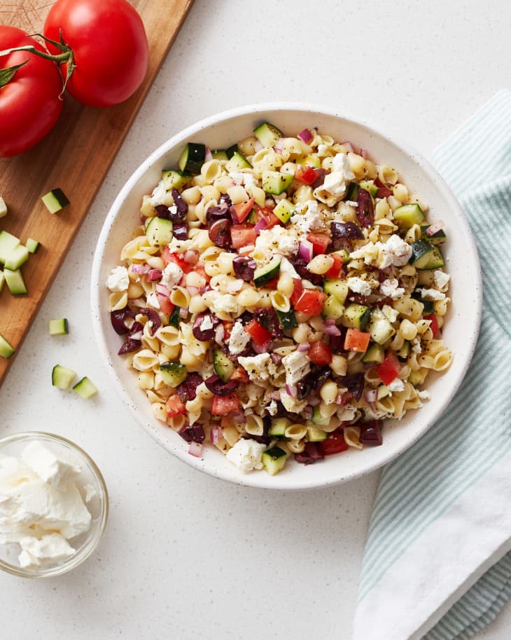 Pasta salad with cucumbers, tomatoes, olives, and feta in a bowl, next to fresh tomatoes and a striped cloth.