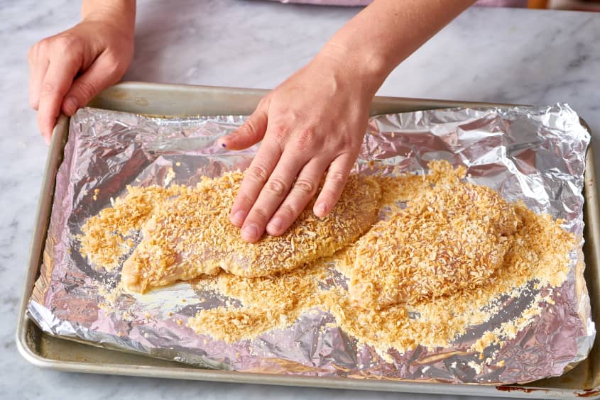 Hands pressing breadcrumbs onto chicken breasts on a foil-lined baking sheet.