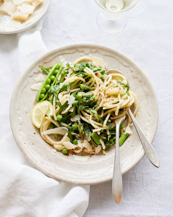 Spaghetti with asparagus, peas, lemon slices, and shaved parmesan on a white plate with a fork and spoon.