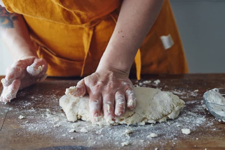 Person kneading dough on a floured wooden surface, wearing an orange apron.