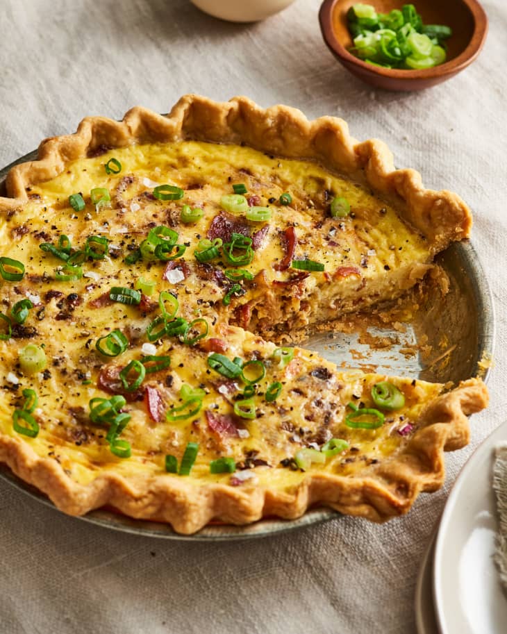 Savory quiche with bacon and green onions in a flaky crust, served on a table with a small bowl of chopped scallions.
