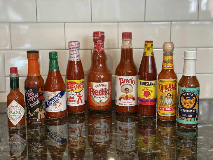 Assorted hot sauce bottles on a kitchen counter with a tiled backsplash.