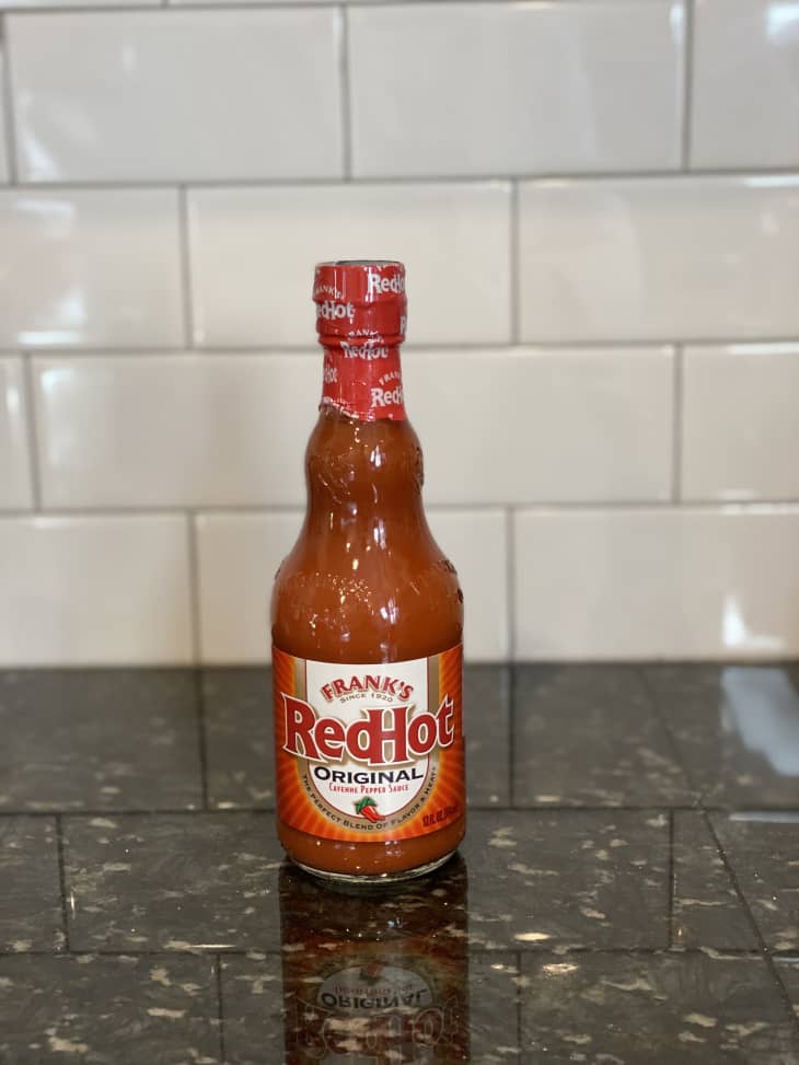 Frank's RedHot Original Cayenne Pepper Sauce bottle on a dark granite countertop with white subway tile backsplash.