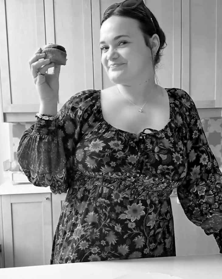Person smiling in a kitchen while holding up a cupcake.