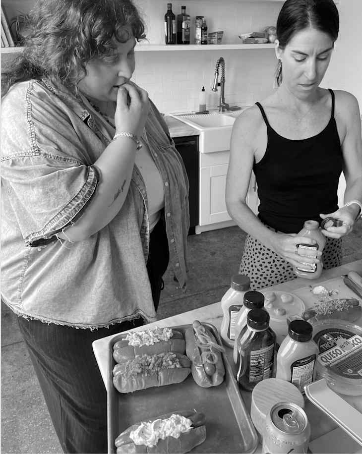 Two people in a kitchen preparing hot dogs at a counter.