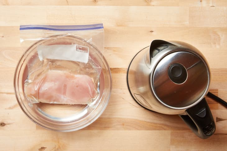 Chicken breast in a sealed bag in a bowl next to a stainless steel kettle on a wooden surface.