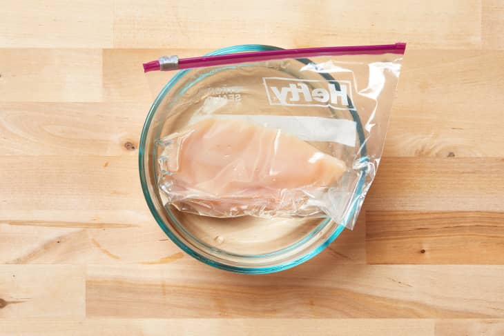 Raw chicken breast in a sealed plastic bag submerged in a glass bowl of water on a wooden surface.