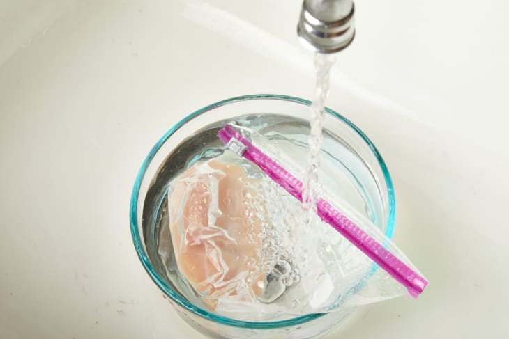 Chicken breast in a sealed bag submerged in a bowl under running water.