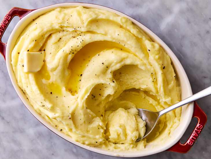 Creamy mashed potatoes in a red baking dish, topped with melted butter and black pepper, with a spoon resting inside.