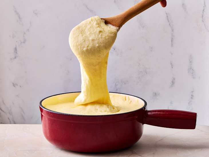 Creamy cheese mixture being stirred with a wooden spoon in a red pot against a light marble background.