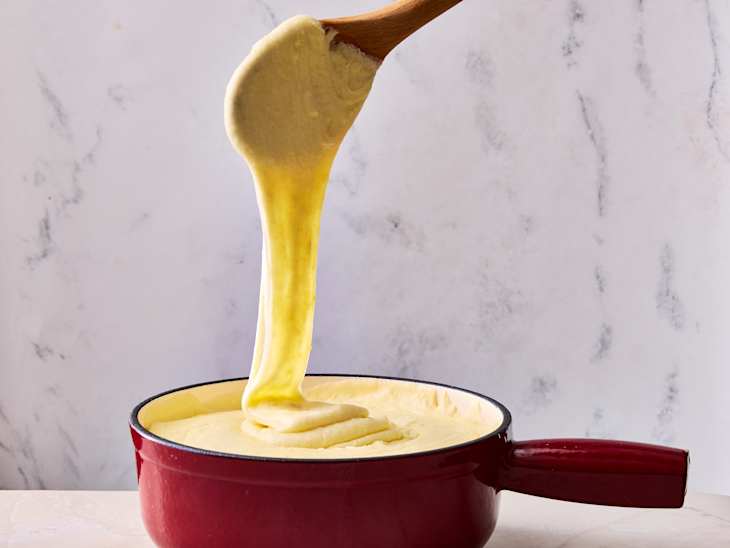 Creamy yellow batter being stirred with a wooden spoon in a red pot against a light marble background.