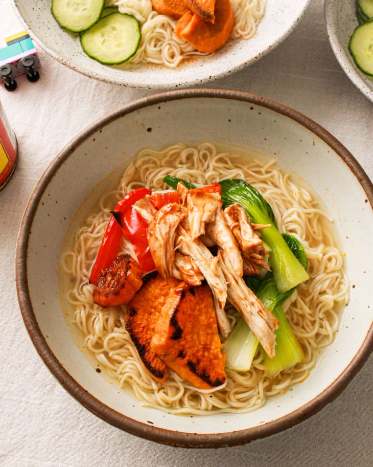 Bowl of ramen with grilled chicken, red peppers, bok choy, and sweet potatoes on a light tablecloth.