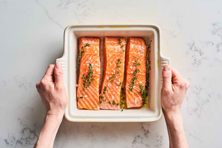 Three salmon fillets with fresh thyme in a baking dish held by two hands.
