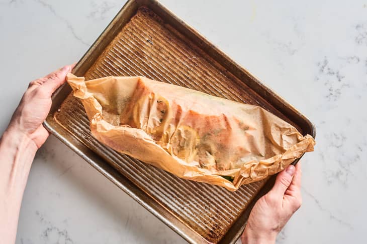 Hands holding a parchment-wrapped dish on a ribbed baking sheet.