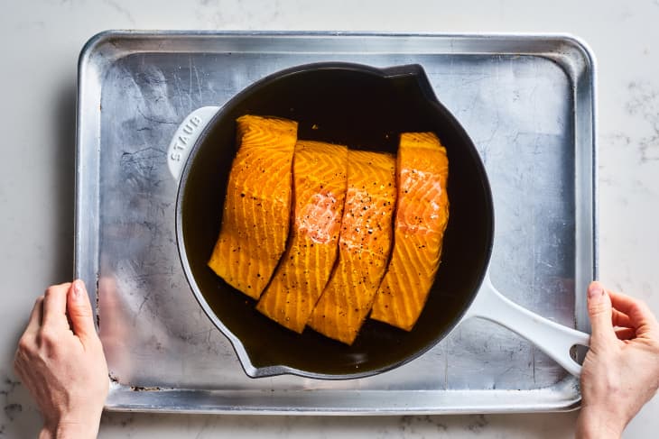 Salmon fillets seasoned with pepper in a white skillet on a metal baking sheet, held by two hands.