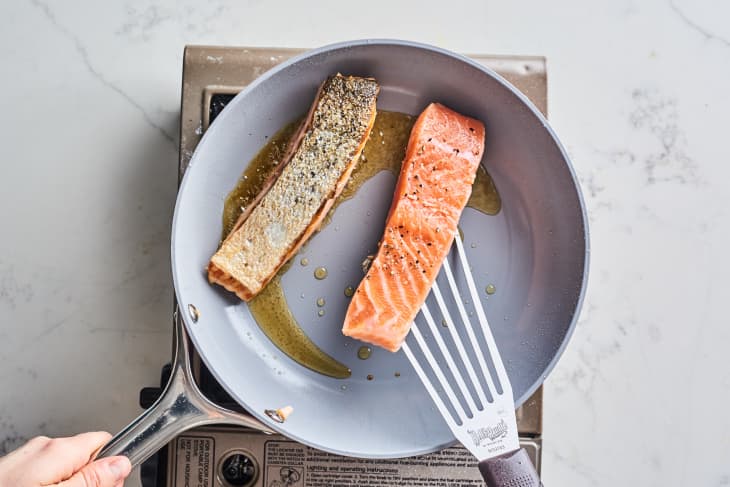 Two salmon fillets cooking in a gray frying pan on a stovetop, one being flipped with a spatula.