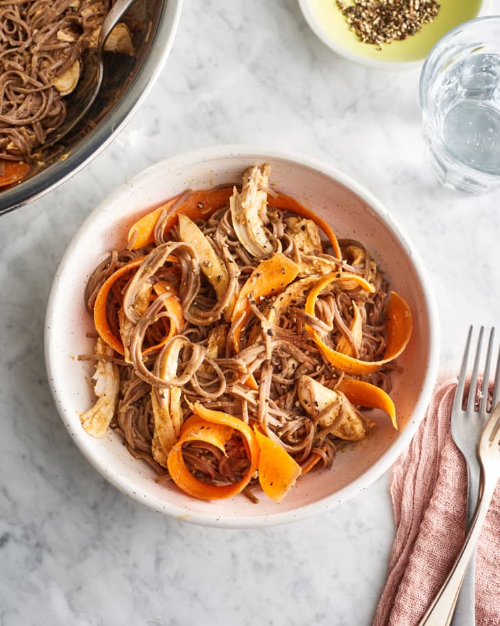 Soba noodles with shredded chicken and carrot ribbons in a bowl on a marble surface, with a fork and napkin nearby.