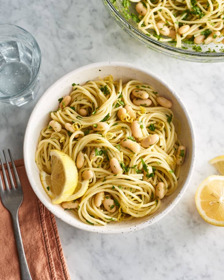 Bowl of cold noodles with white beans, lemon slices, and herbs on a marble surface.